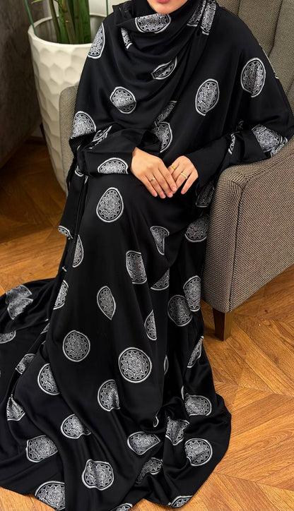 Sitzende Frau im schwarzen Abaya Set Mahira mit silberfarbenen Ornamenten und weit fallendem Butterfly-Schnitt.